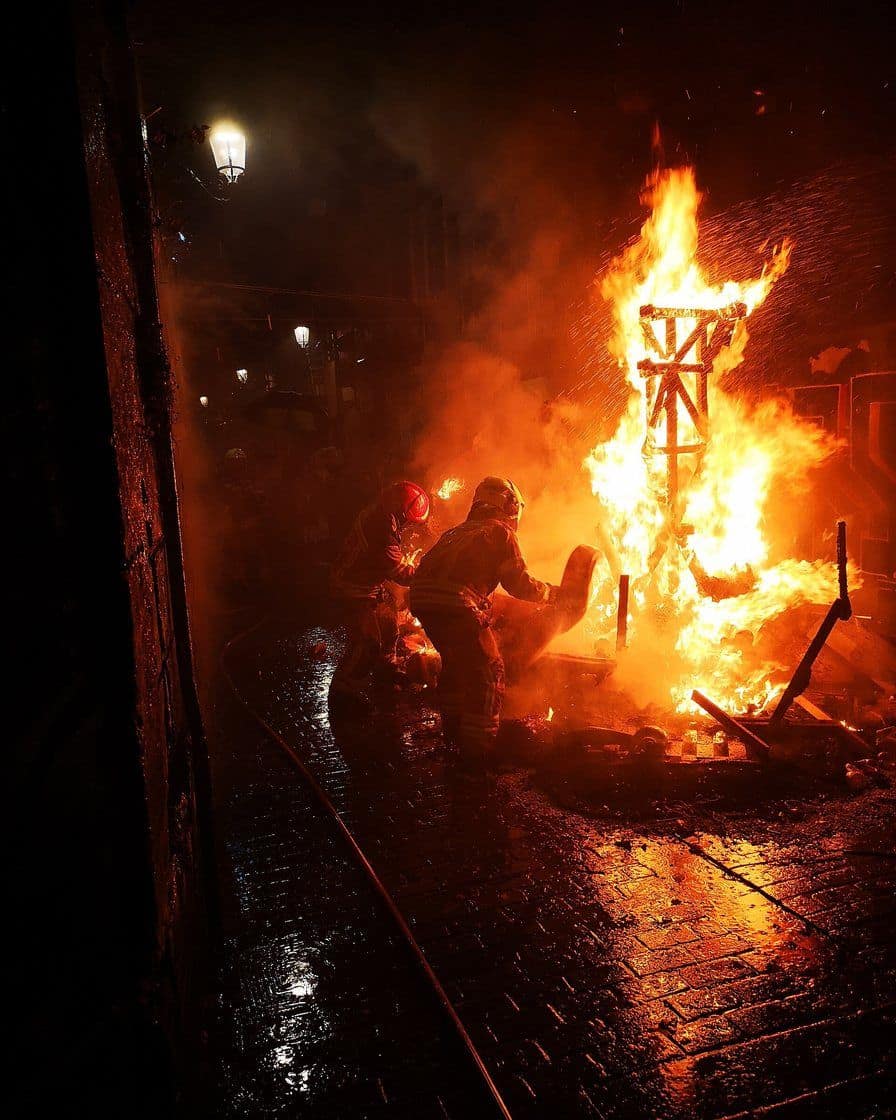 Cremà de una falla en una calle de Valencia de noche, monumento en llamas con humo denso y bomberos vigilando junto a la valla