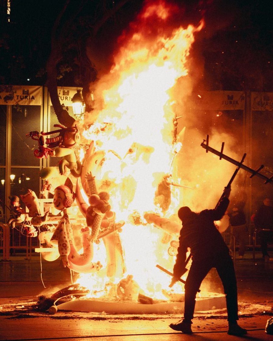 Falla grande ardiendo de noche en una calle de Valencia, con las llamas iluminando las fachadas y el público detrás de las vallas