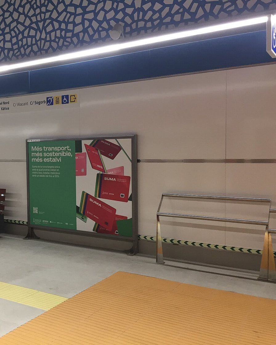 Platform of a Metrovalencia station with a train pulling in, passengers waiting behind the yellow line and station signs in the background